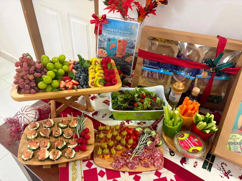 A fruit platter of grapes, berries and kiwi beside smoked salmon cucumber bites, olive and salami skewers, salad, crudités and a gift box